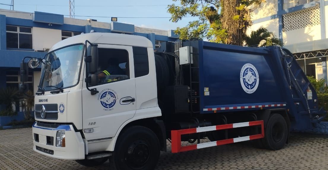 Camión de basura, cabina blanca, contenedor azul, estacionado frente a un edificio azul, de día.