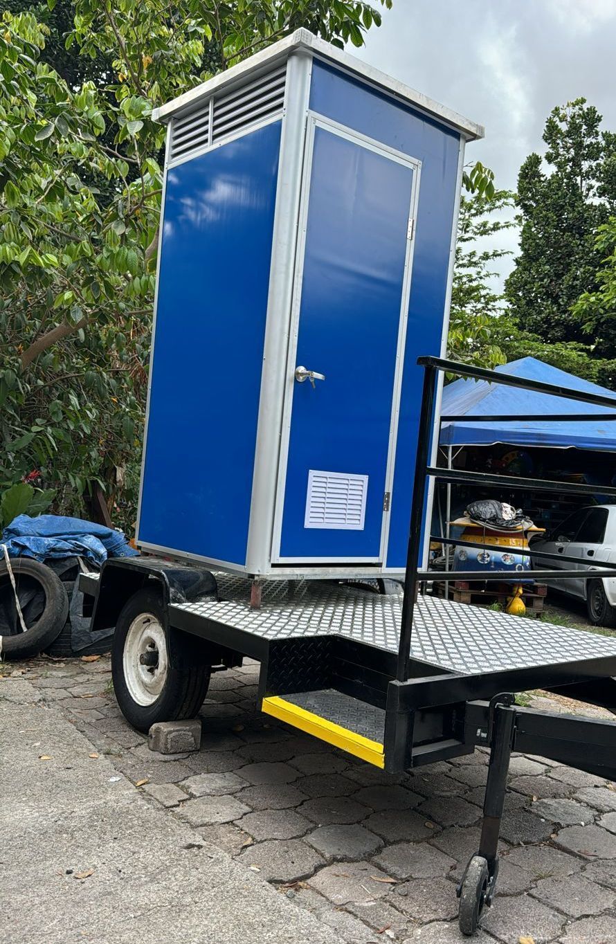 Baño portátil azul sobre un remolque con estructura y puerta metálicas, estacionado al aire libre.