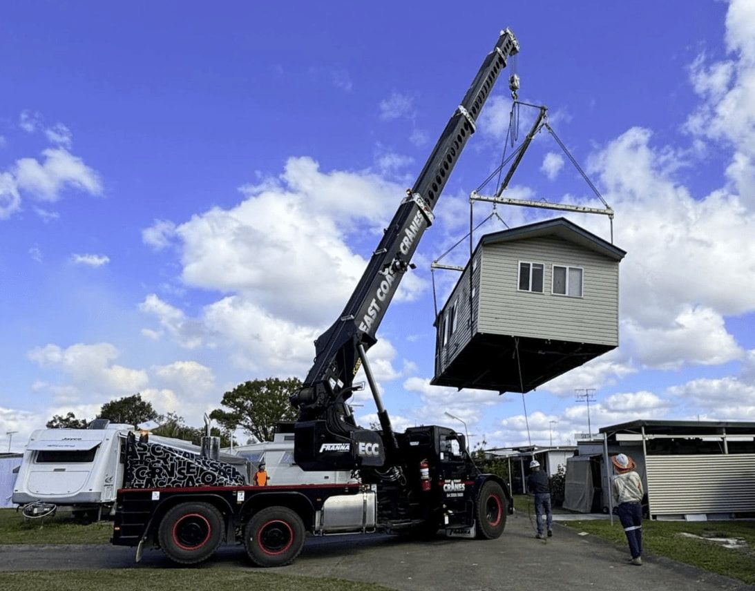 A Crane Lifting a Small House on a Truck Bed in a Field on a Sunny Day — East Coast Cranes in Newcastle, NSW