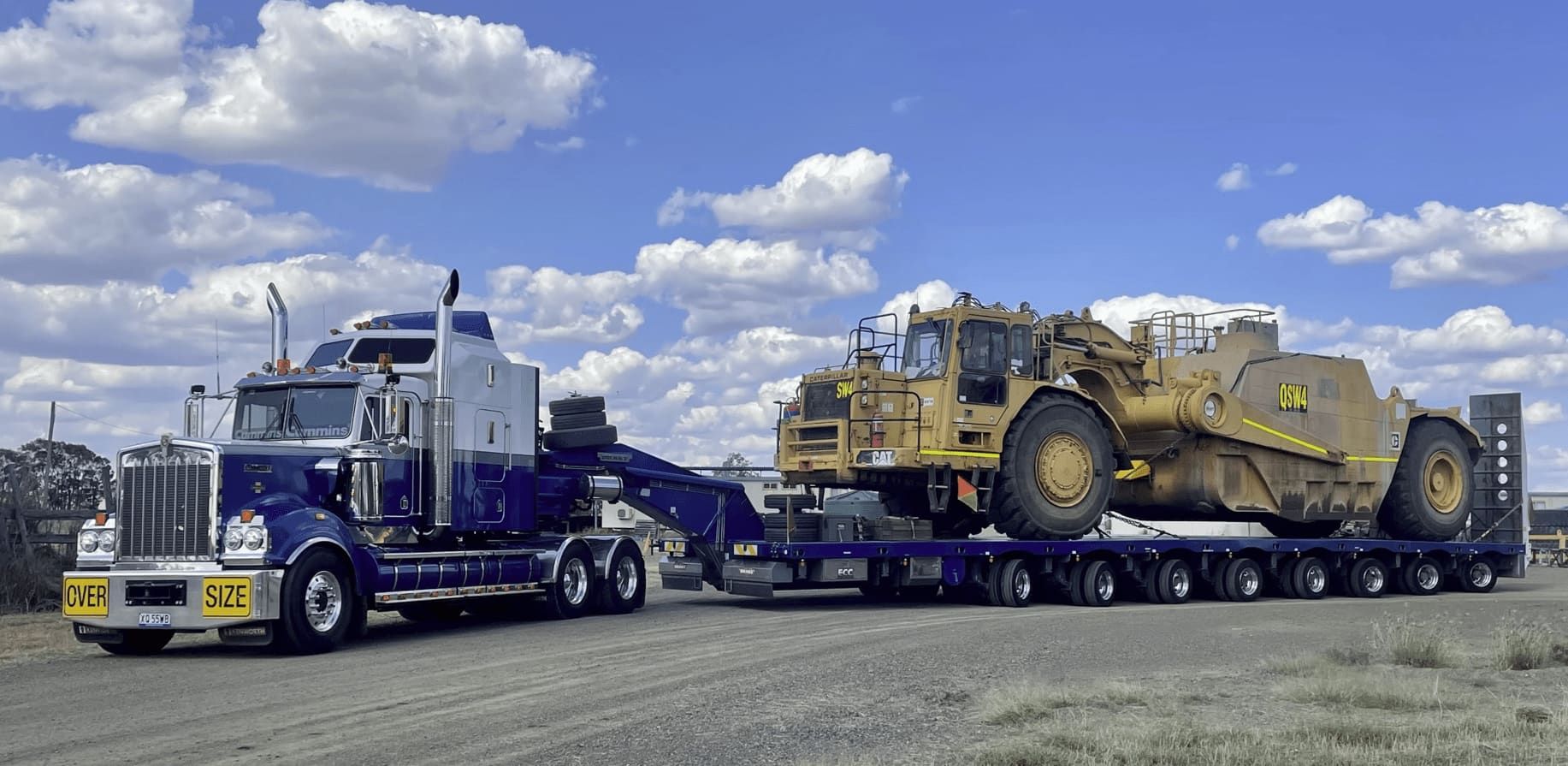 Blue Semi-truck Hauling a Large Yellow Earth Scraper on a Flatbed Trailer Under a Partly Cloudy Sky — East Coast Cranes in Coffs Harbour, NSW