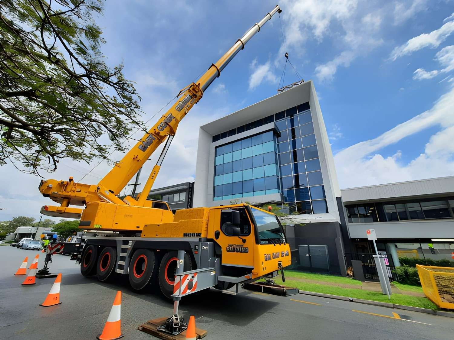 Yellow Crane Lifting at a Modern Building With Glass Windows, Cones on the Road, Cloudy Sky — East Coast Cranes in Coffs Harbour, NSW