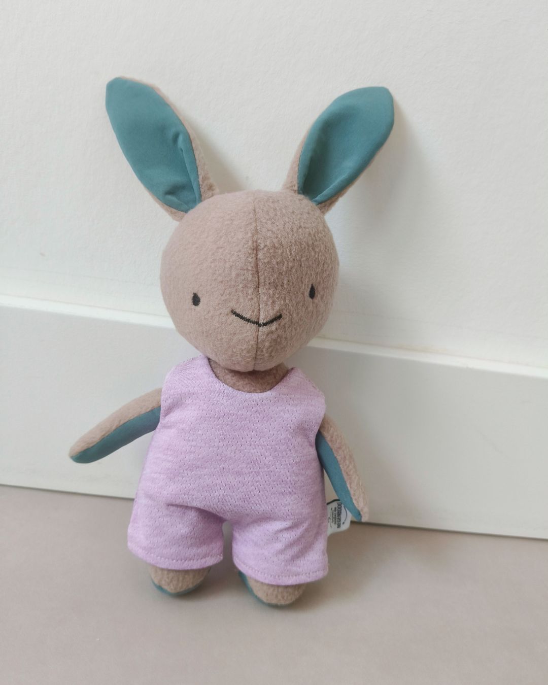 Klein pluche konijntje met blauwe oren en een paars pakje, staand tegen een witte muur. van kleding overledene