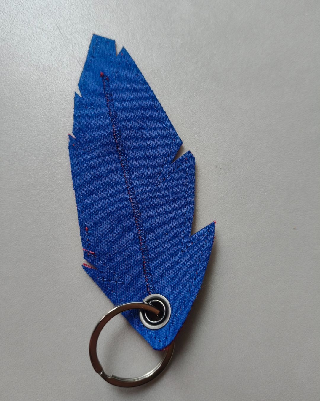 Blauwe sleutelhanger in de vorm van een veer met een metalen ring op een lichtgrijze achtergrond.