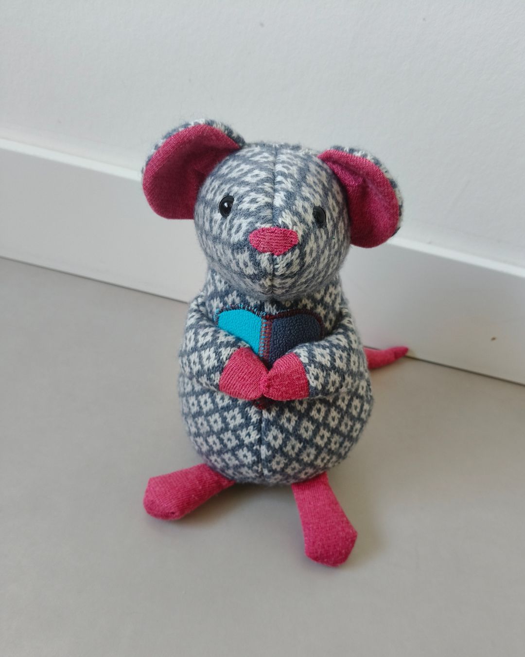 Een roze en grijze gebreide muis van dierbare kleding staat op de vloer tegen een witte muur.