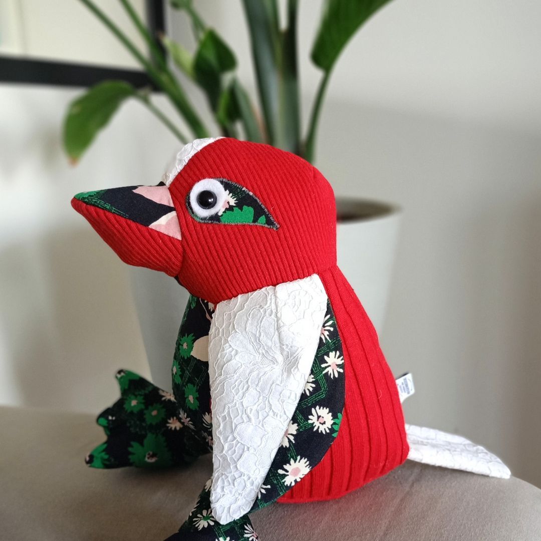 Rood-wit vogeltje met groene bloemaccenten gemaakt van kleding , zittend op een oppervlak binnenshuis.