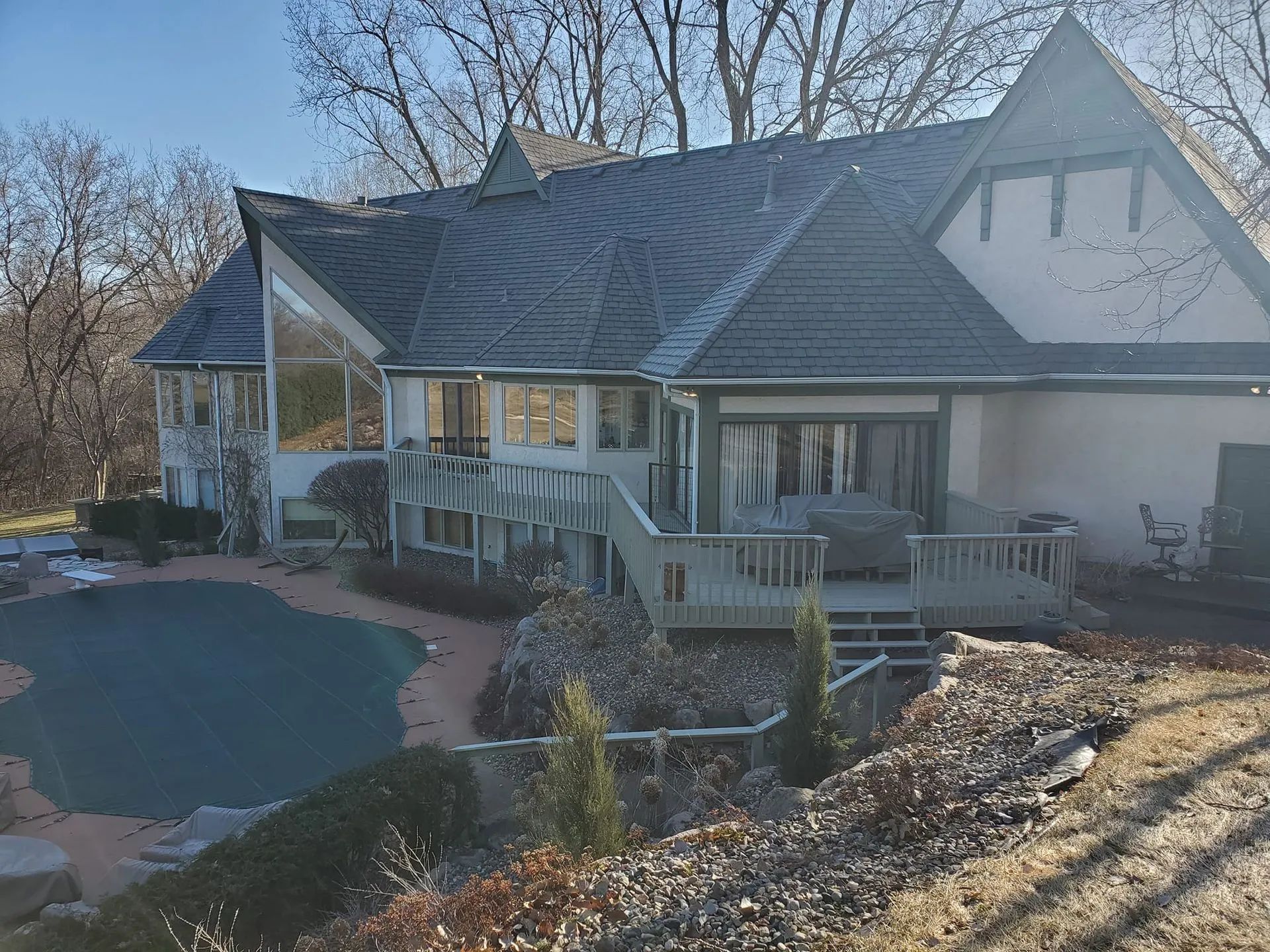 House with a wraparound deck beside a backyard pool, with bare trees and winter sunlight.