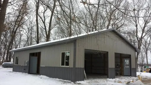 Metal garage building with gray and silver siding, snowy ground, and bare trees.