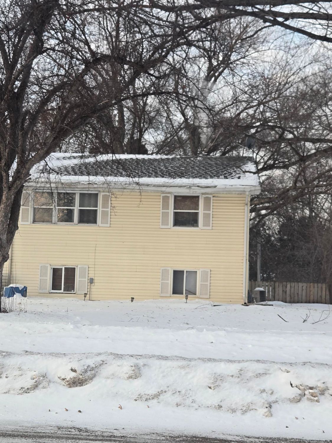 Two-story yellow house with white shutters in a snowy setting. Bare trees in background.