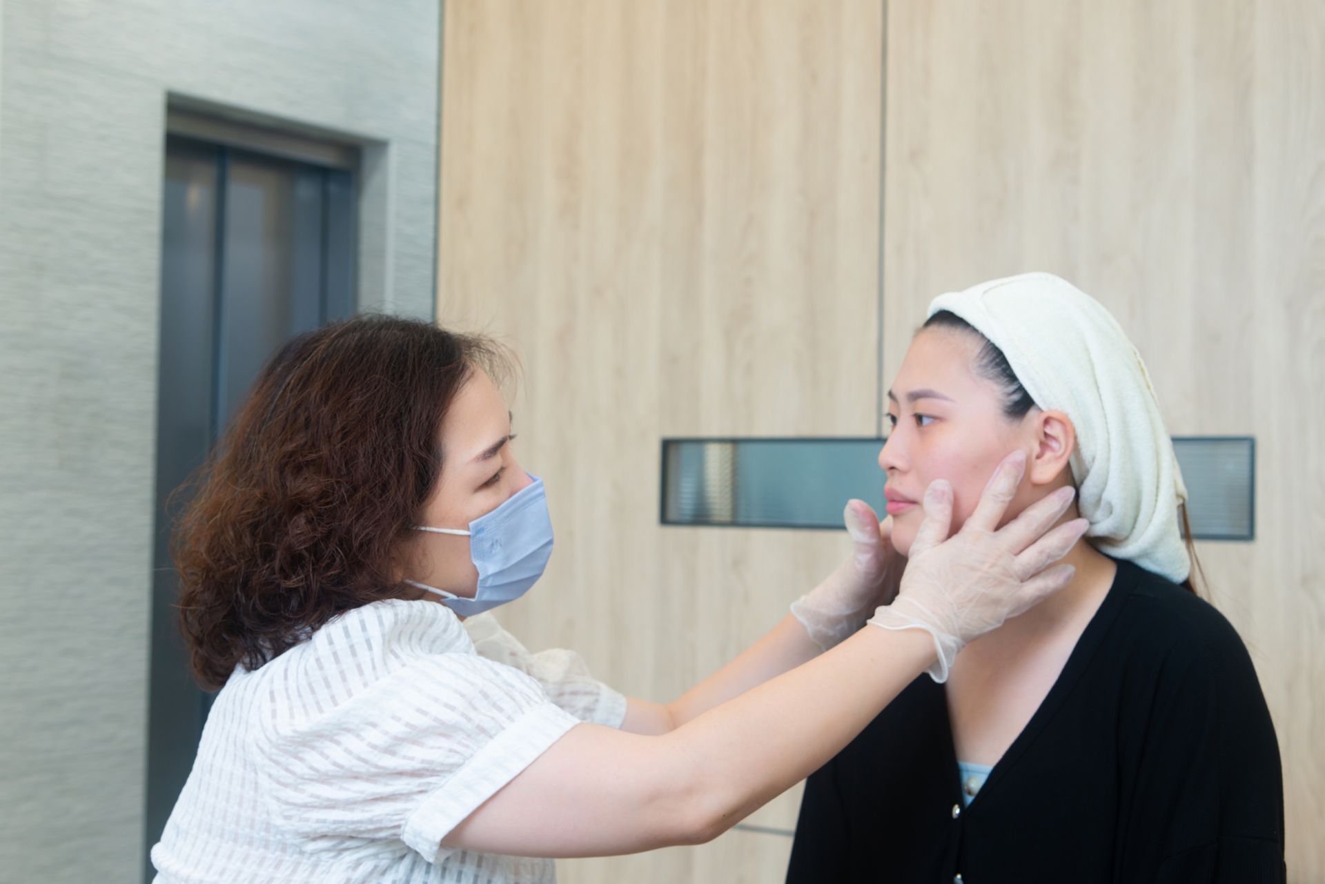 兩名戴著口罩的女子在室內面對面站著；其中一人幫另一人調整口罩和下巴帶。