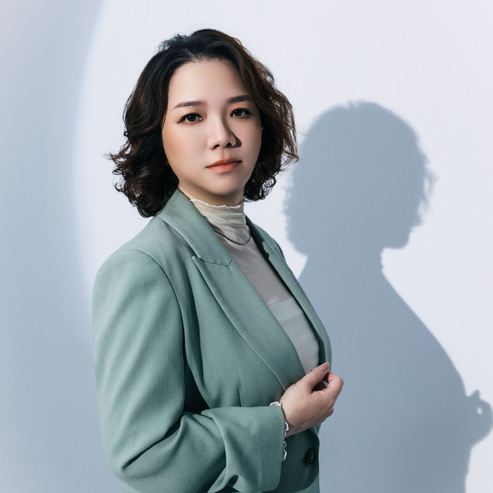 一位身穿薄荷綠西裝外套的女子靠在淺色牆面上，身後投下柔和的陰影。