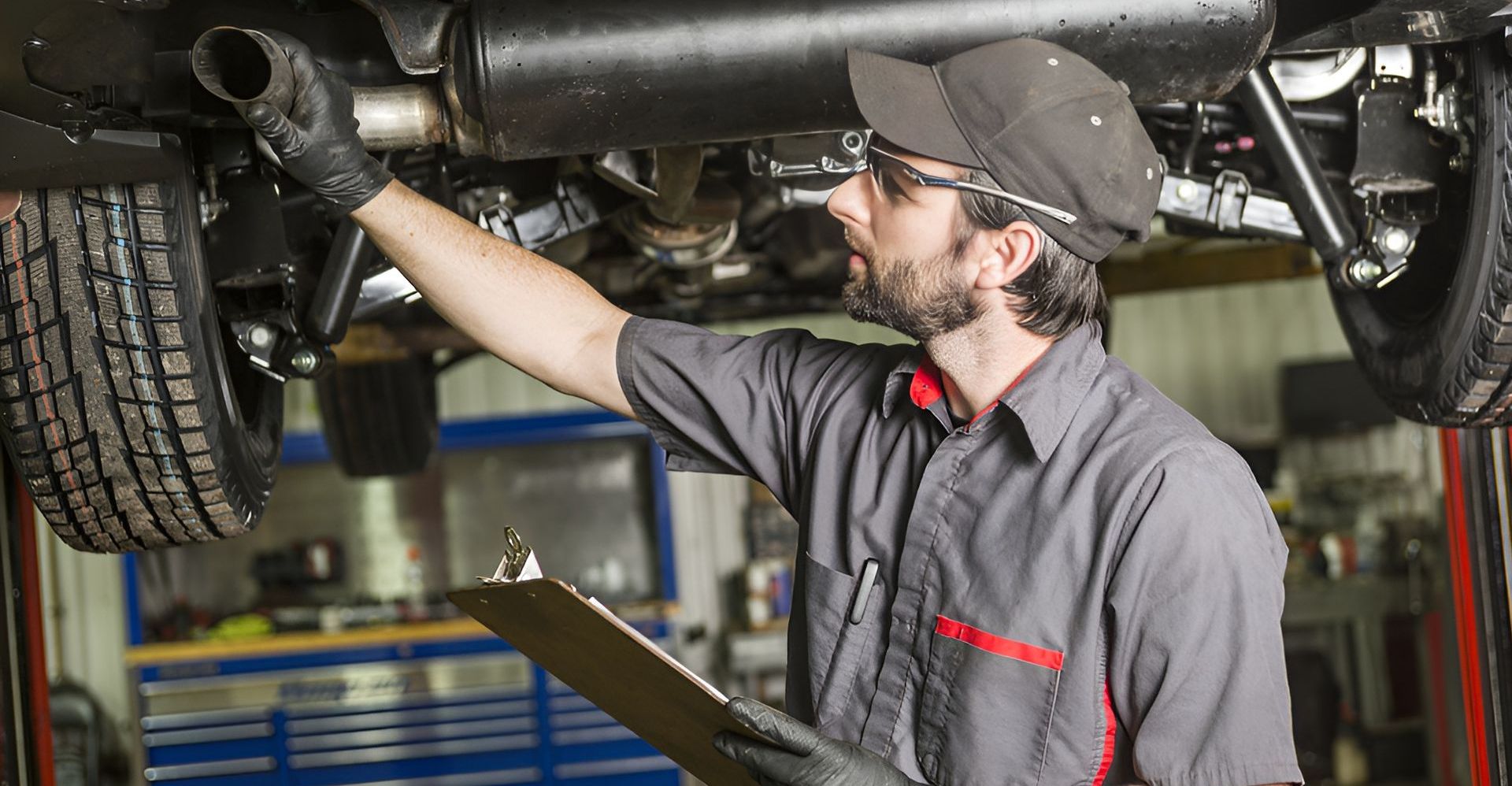 A mechanic wearing glasses, a cap and gloves inspects a car's underside while holding a clipboard.