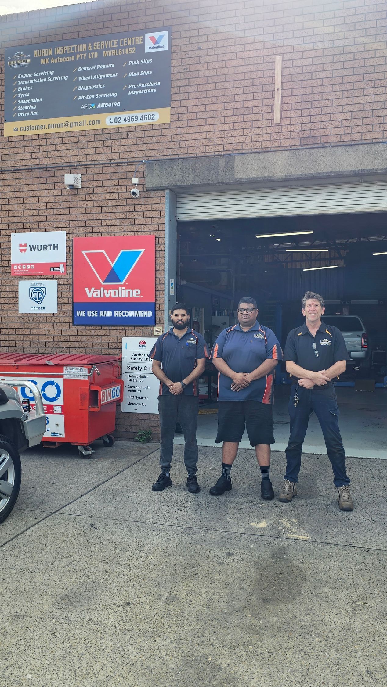 Three men stand in front of an auto repair shop. Valvoline sign visible. Garage door open.