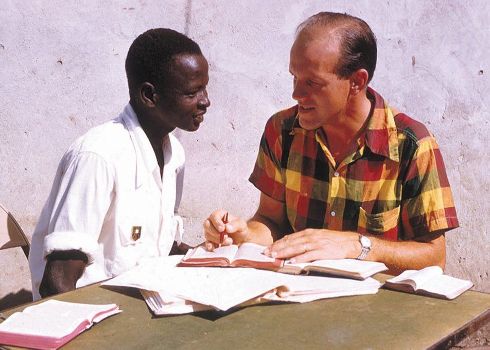 Two people in conversation at a table with papers and books, one in a white coat and one in a plaid shirt.