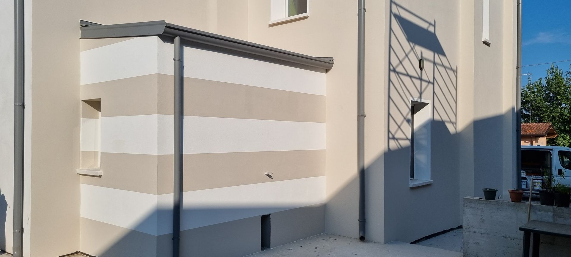 Esterno di un edificio bianco con tenda da sole a strisce, grata alla finestra montata a parete e un passaggio pedonale illuminato dal sole.