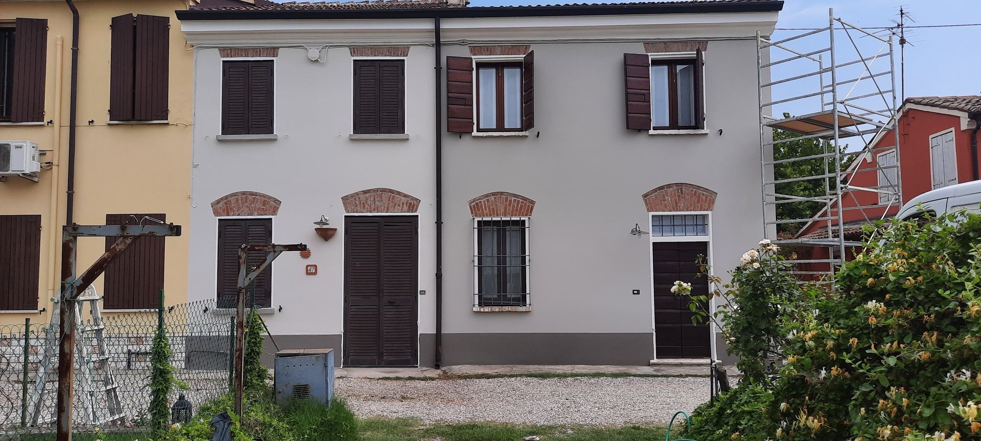 Una fila di case bianche con persiane e porte marroni dietro un basso muro di pietra e vegetazione.