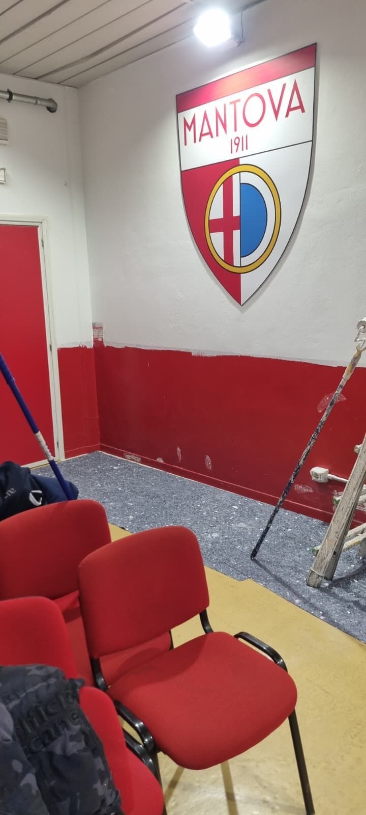 Stanza rossa e bianca con sedie rosse e lo stemma di una società sportiva del Mantova appeso alla parete.