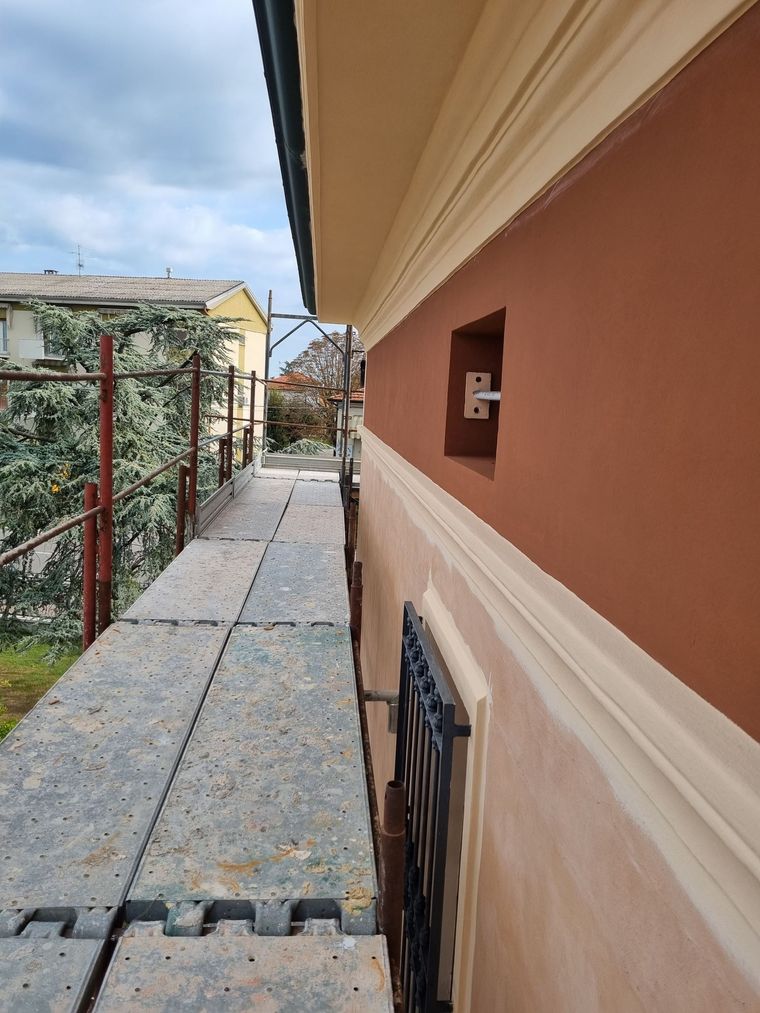 Stretto passaggio pedonale in cemento accanto a una serra con una recinzione rossa e vegetazione sulla sinistra.