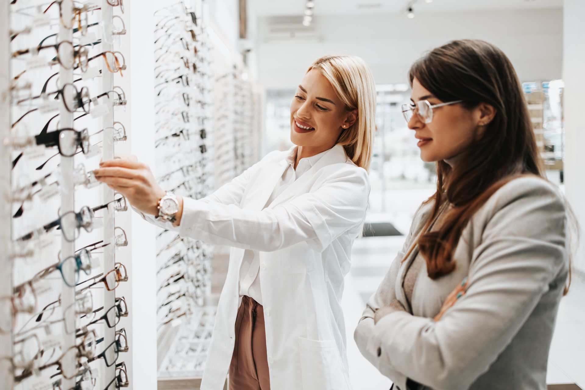 Optician helps customer choose eyeglasses from a display wall in optical store.