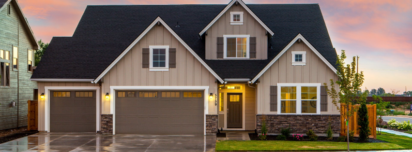 Modern gray suburban house with three garage doors, warm porch light, and landscaped front yard at sunset