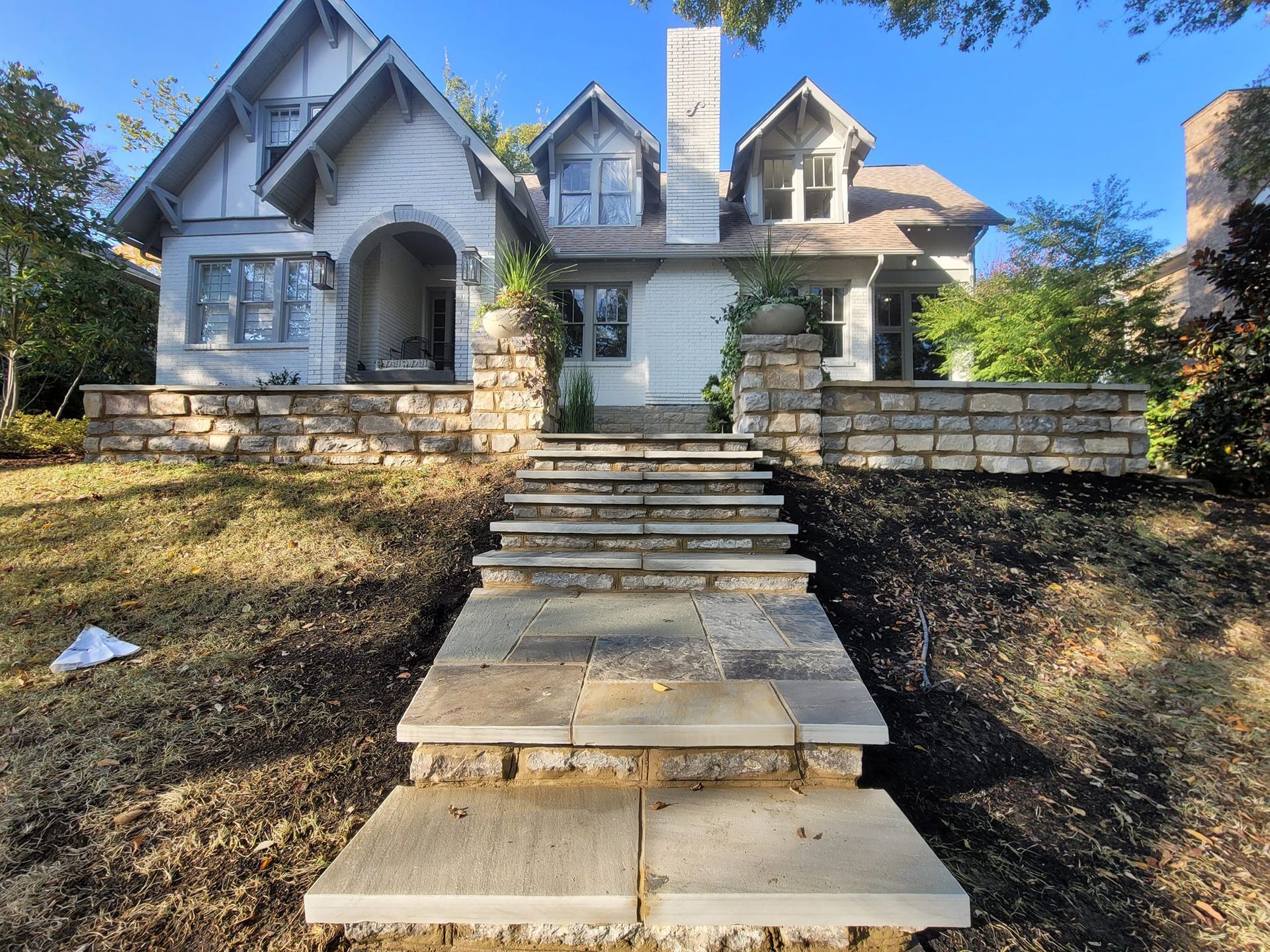 Stone steps lead up to a white house with a stone facade, under a clear, blue sky.