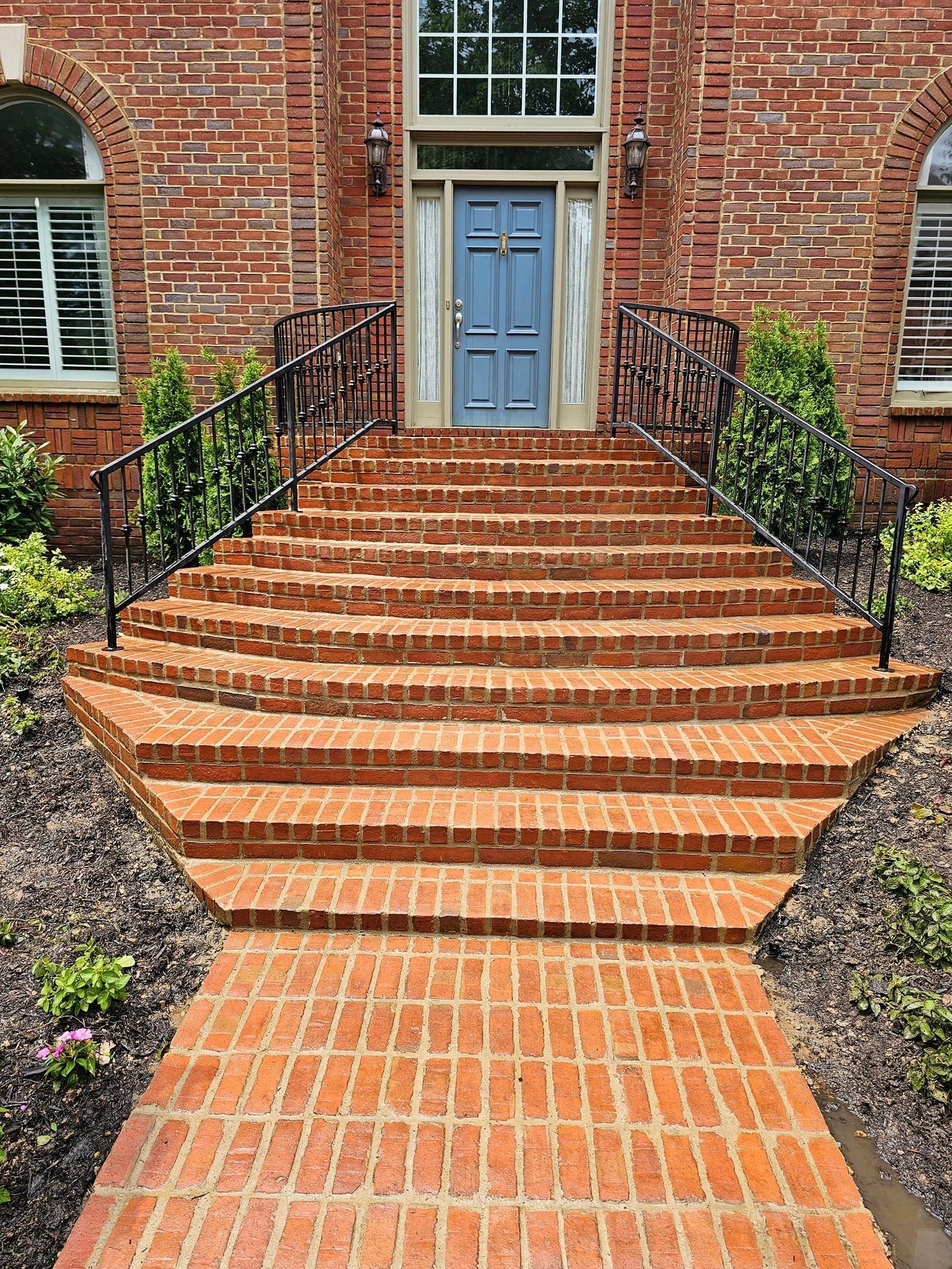 Brick steps lead to a blue door with sidelights, flanked by black railings, shrubs, and a brick walkway.