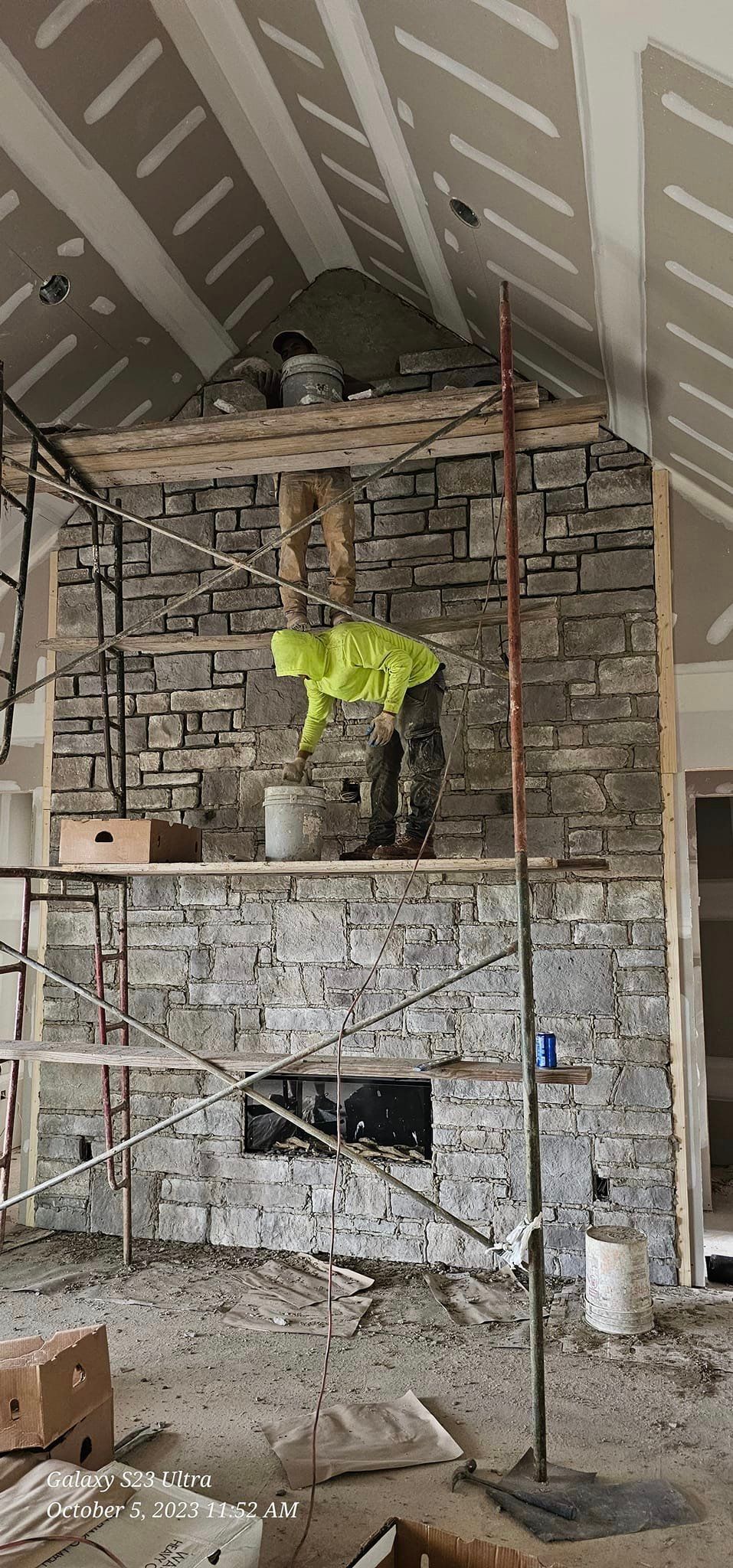 A person in a yellow vest is working on a stone fireplace, with scaffolding and construction materials.