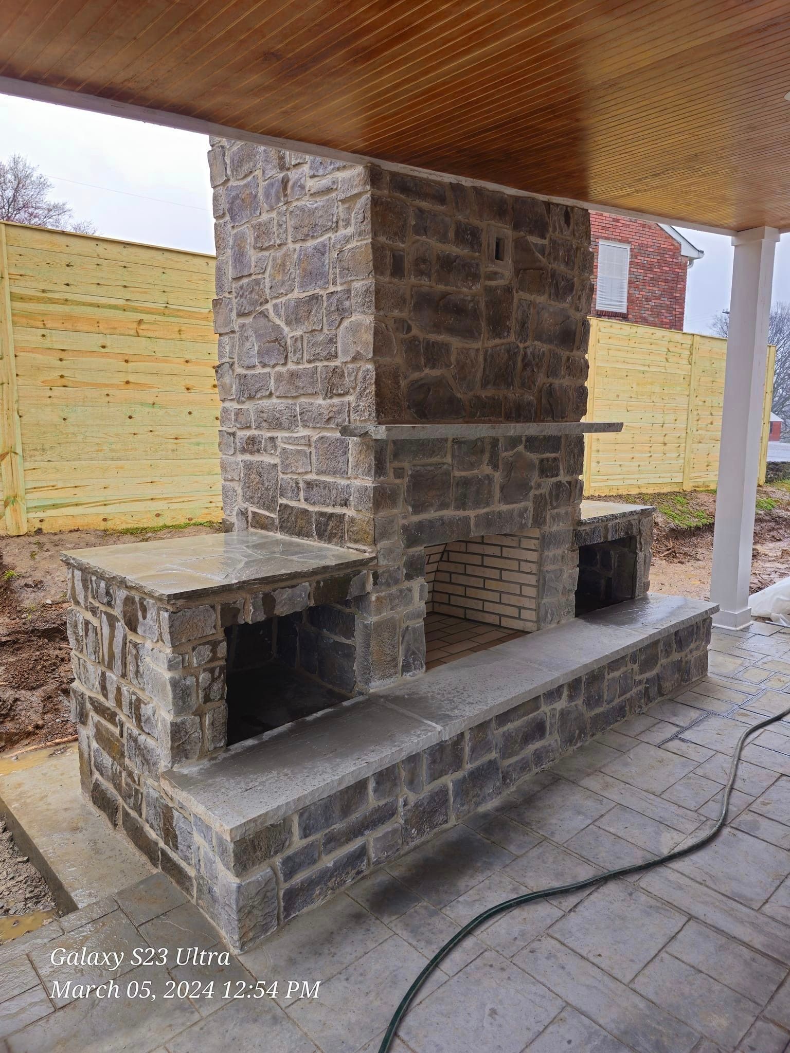 Stone outdoor fireplace with a wood ceiling, set on a concrete patio, with a wood fence in the background.