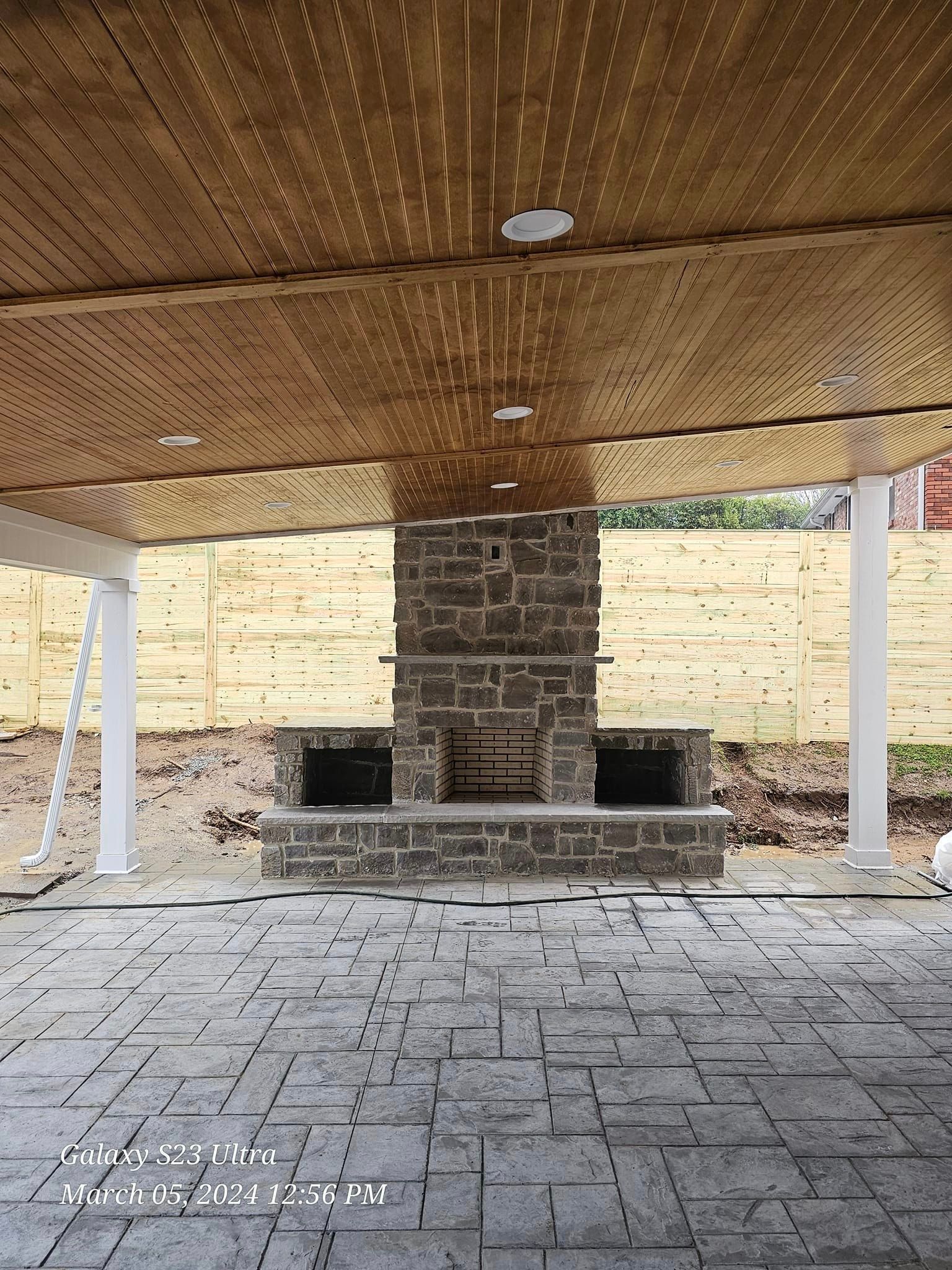 Stone fireplace on a patio with wood ceiling, white pillars, and a wooden fence.