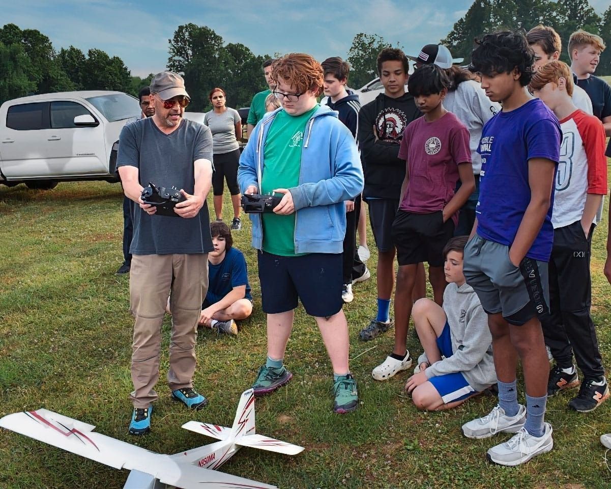 Man and student flying model airplane outdoors with group watching.
