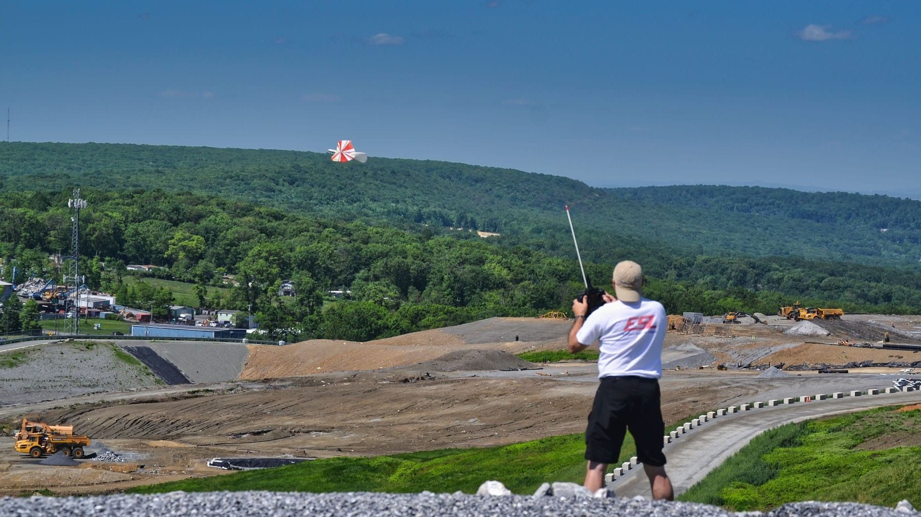 Man flying a kite near a construction site, green hills in the background. Sunny day.
