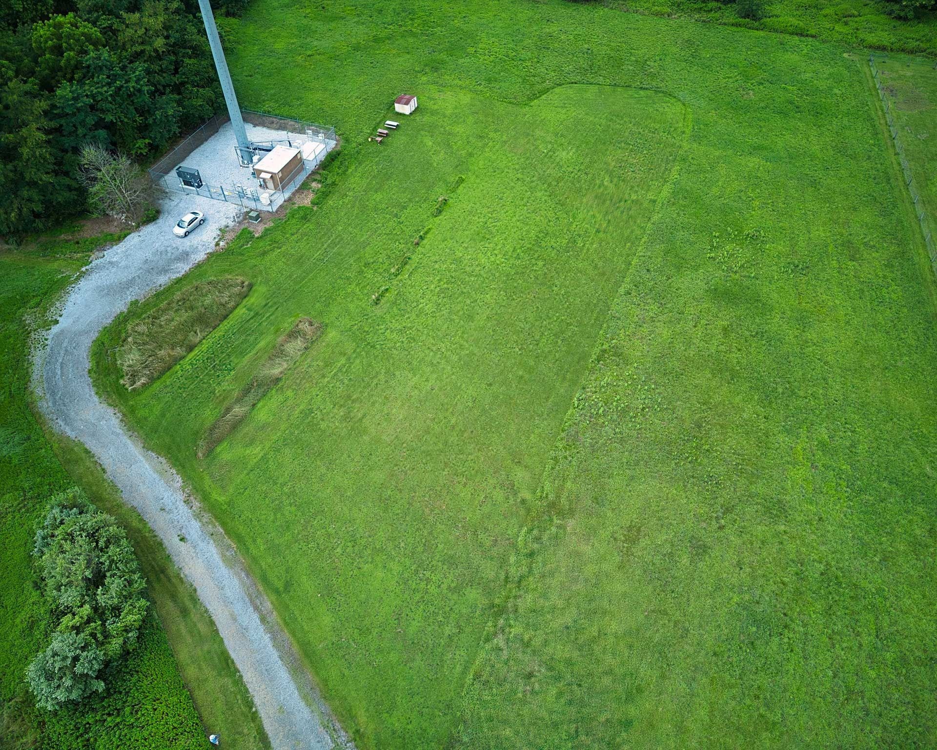 Aerial view of a green field with a gravel road, communication tower, and small buildings.