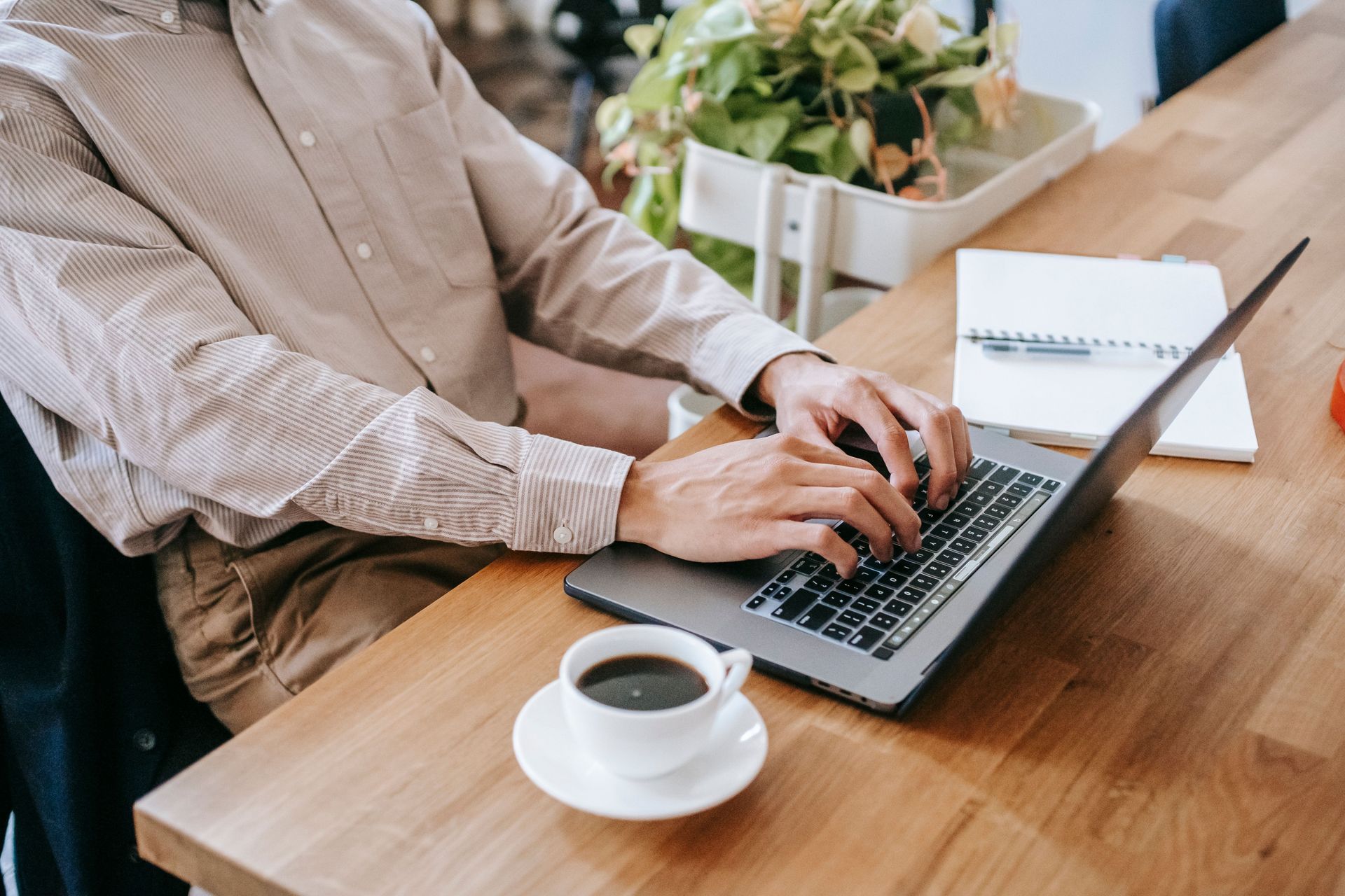 Person typing on a laptop at a wooden table beside a cup of coffee and a small potted plant.