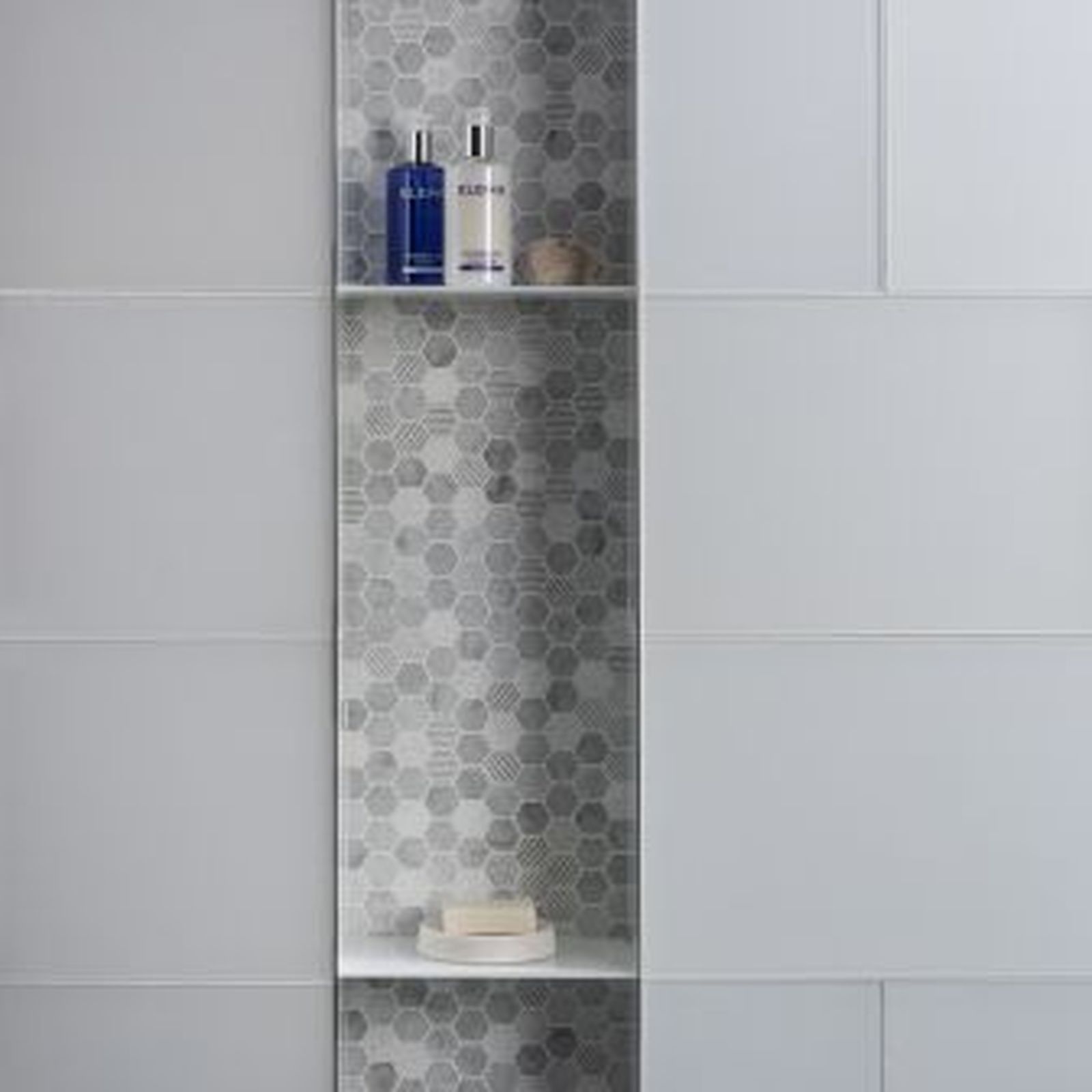 Built-in shower niche with gray hexagon tile and shelves holding toiletries. White rectangular tiles surround the niche.