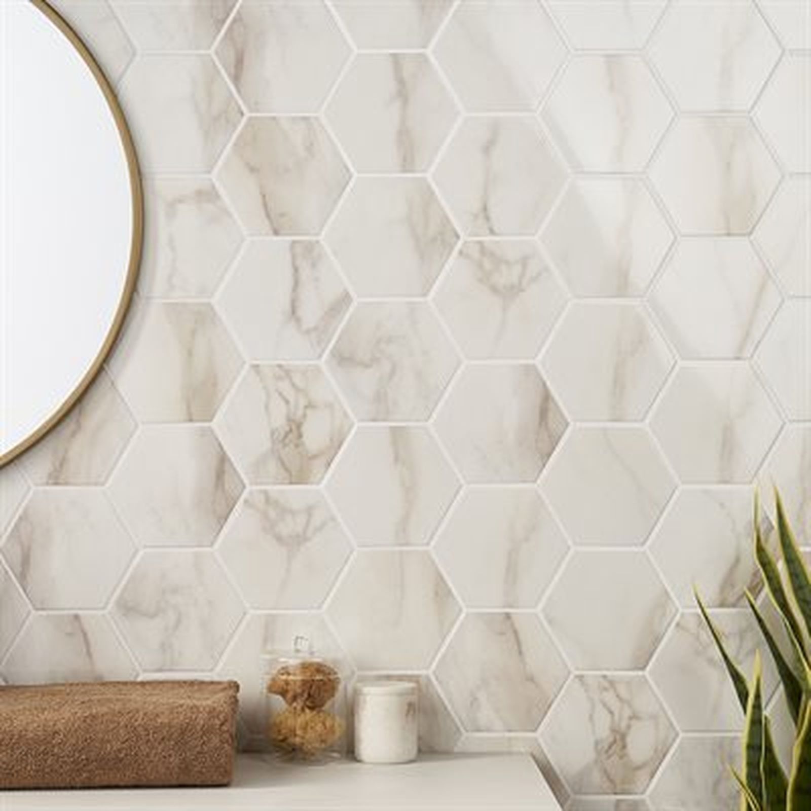 White and tan marble hexagon tiles on a bathroom wall, with a gold-rimmed mirror, brown towel, and plant.