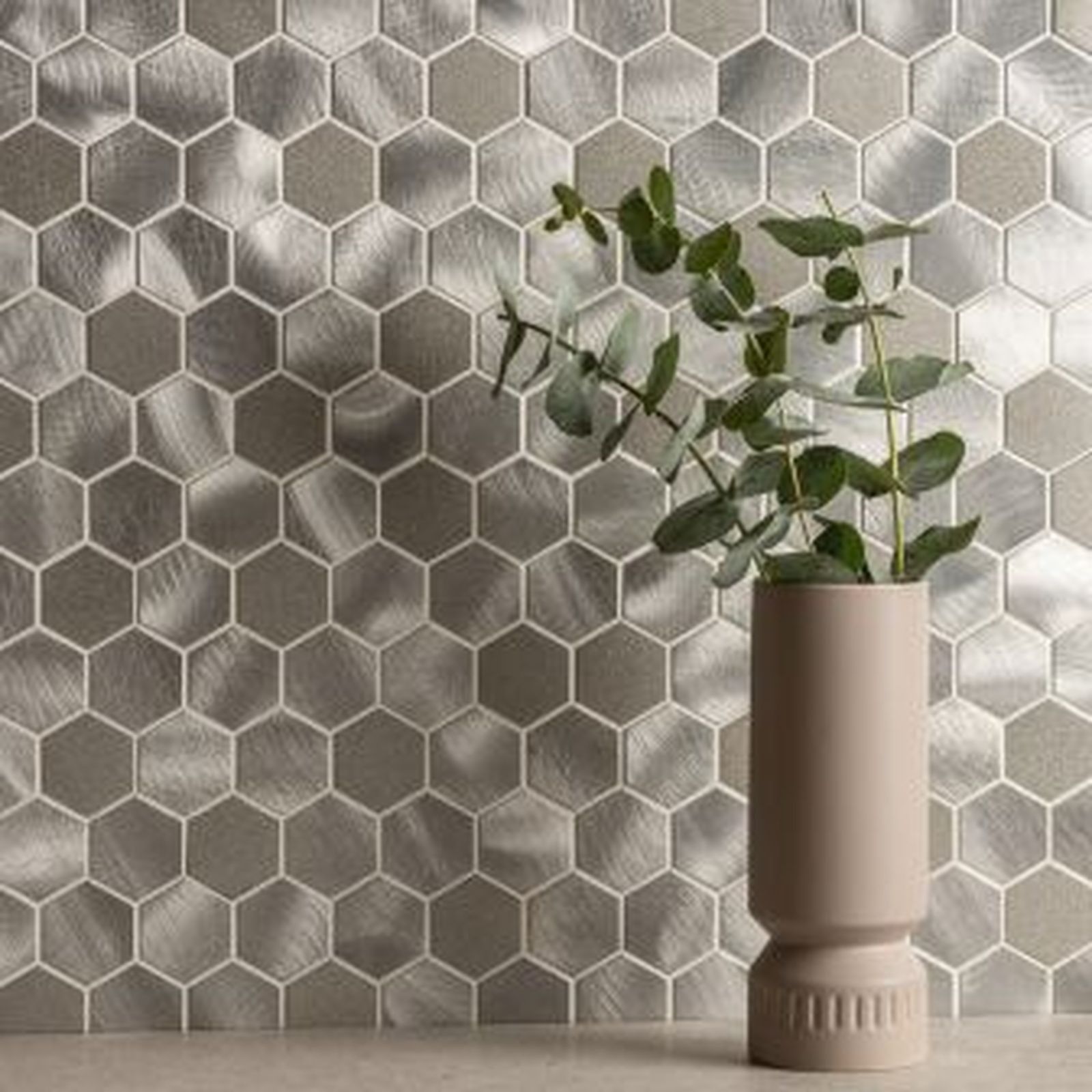 Silver, hexagon-shaped tiles on a wall with a vase holding a sprig of green eucalyptus.