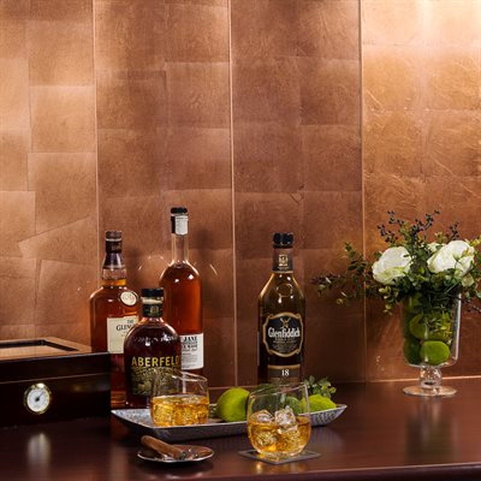 Bar with bottles, glasses of whiskey, a humidor, and a floral arrangement set against a copper-tiled wall.