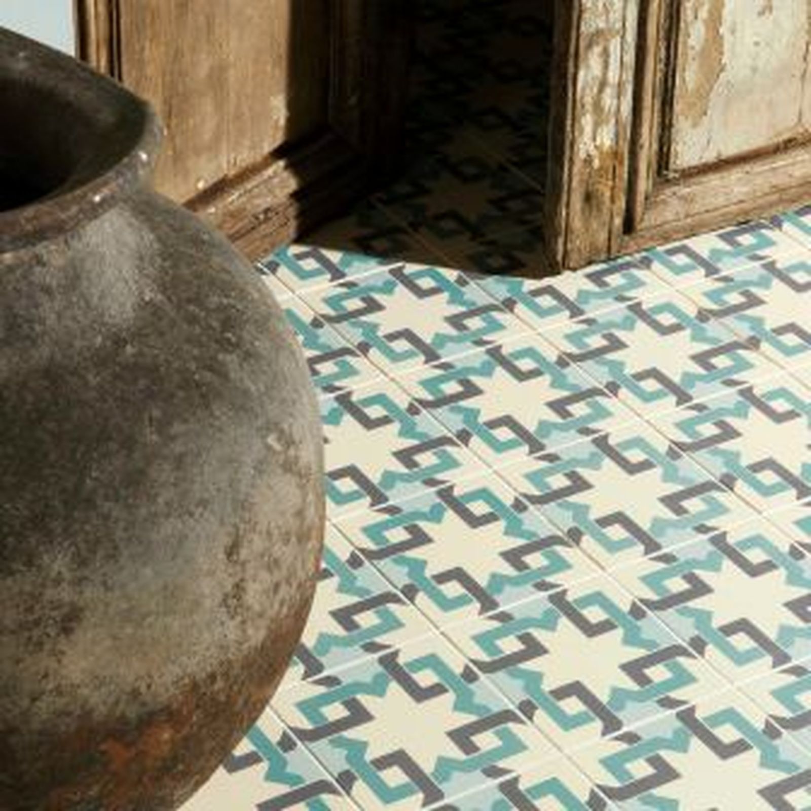 Antique ceramic vase next to a tiled floor with geometric turquoise and gray patterns, near a weathered door frame.