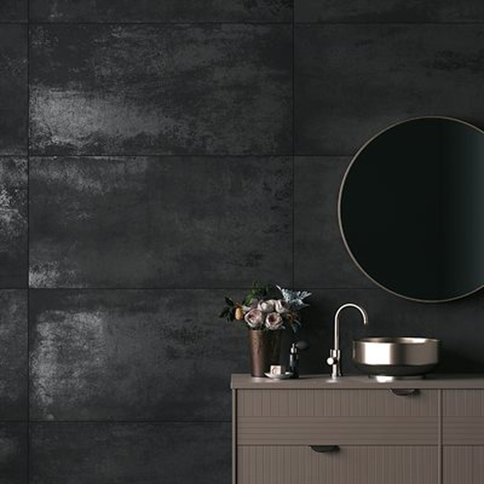Bathroom with dark metallic-look tile walls, round mirror, wooden cabinet with a sink, and a flower arrangement.
