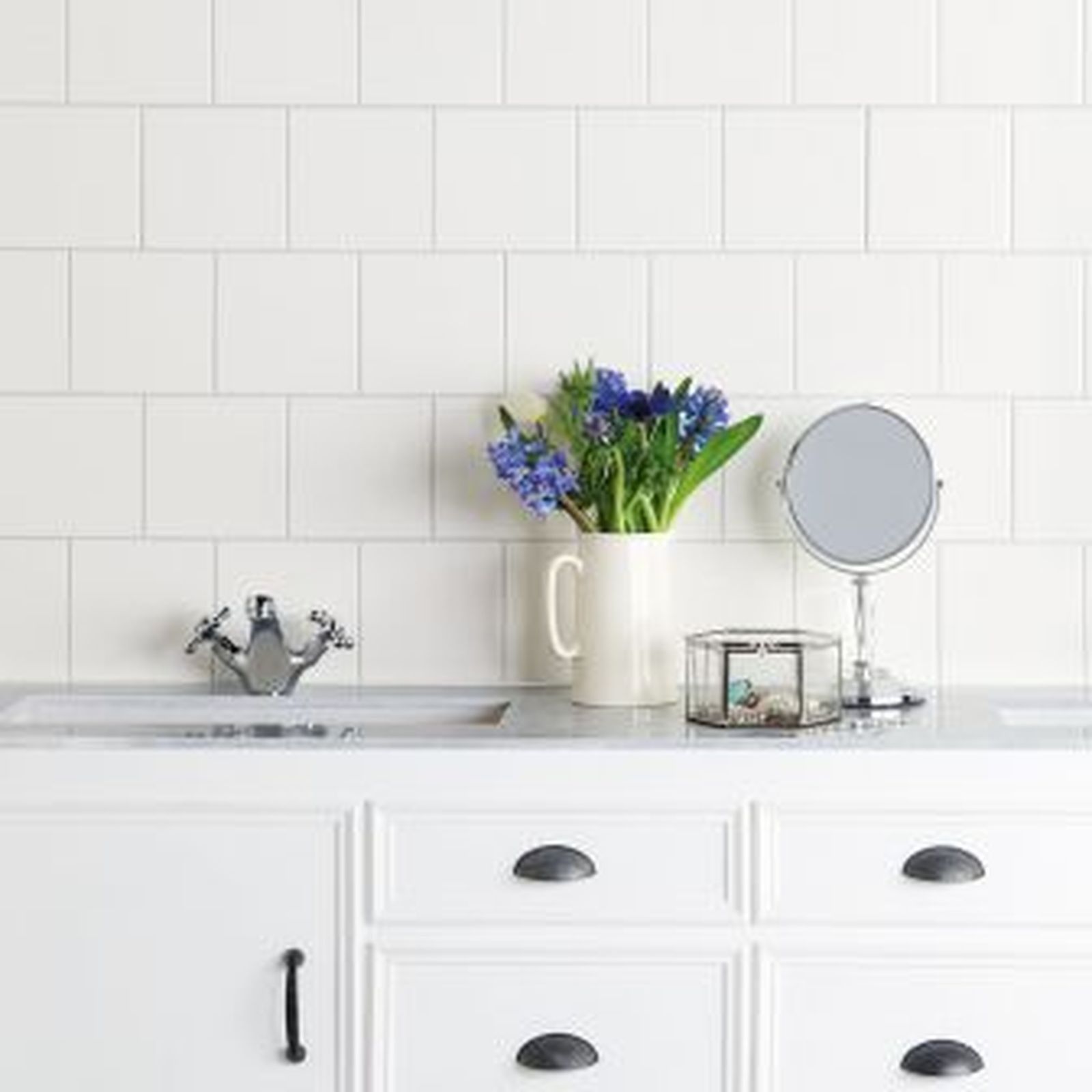 White bathroom vanity with sink, flowers, mirror, and white tiled wall.