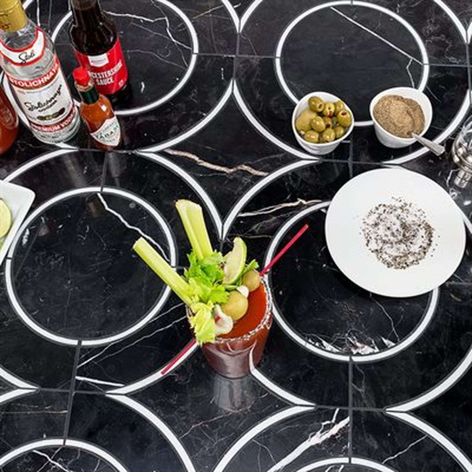 Bloody Mary cocktail on a black and white tiled surface, with ingredients and garnishes.
