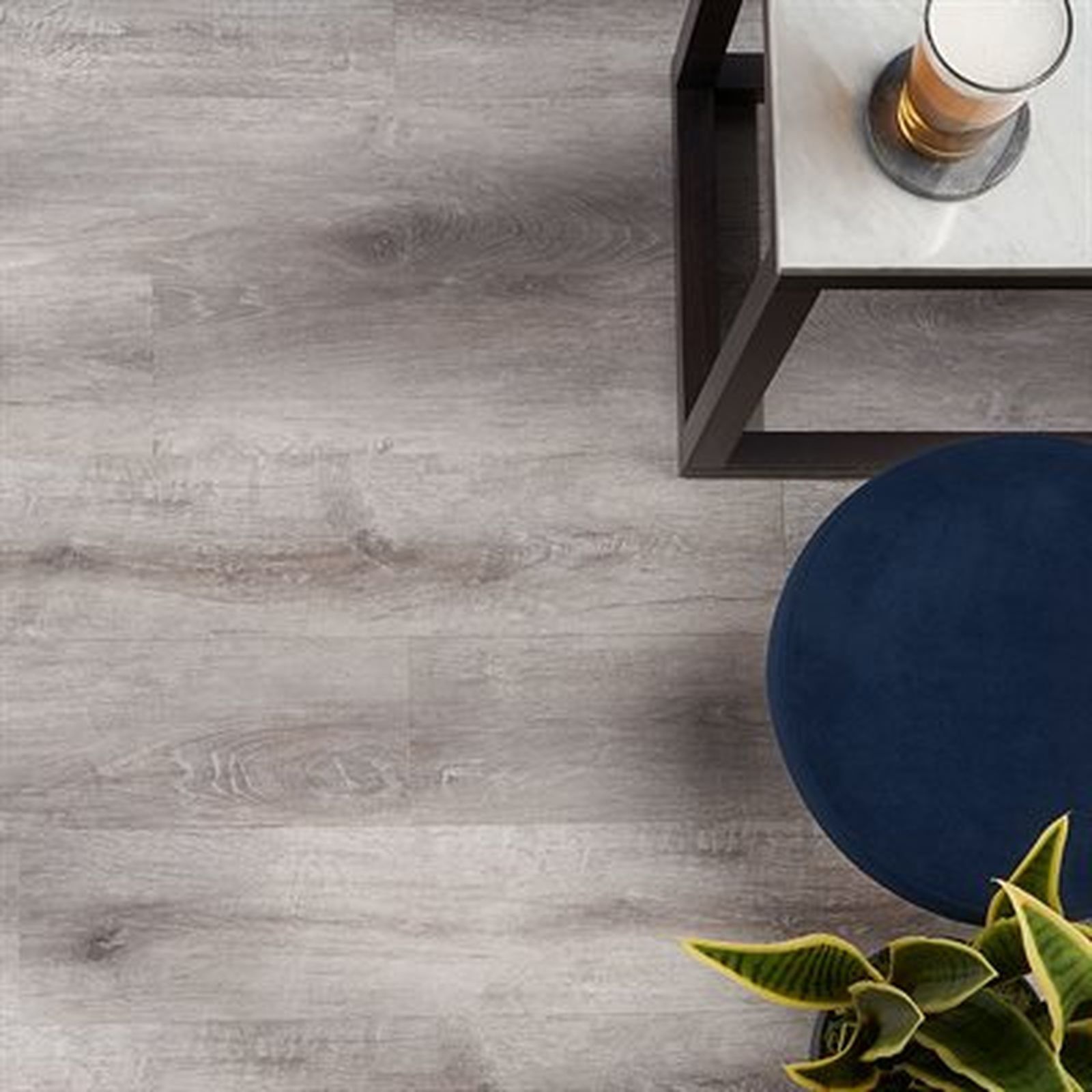 Grey wood-look floor with a small table, a drink, a blue stool, and a plant.