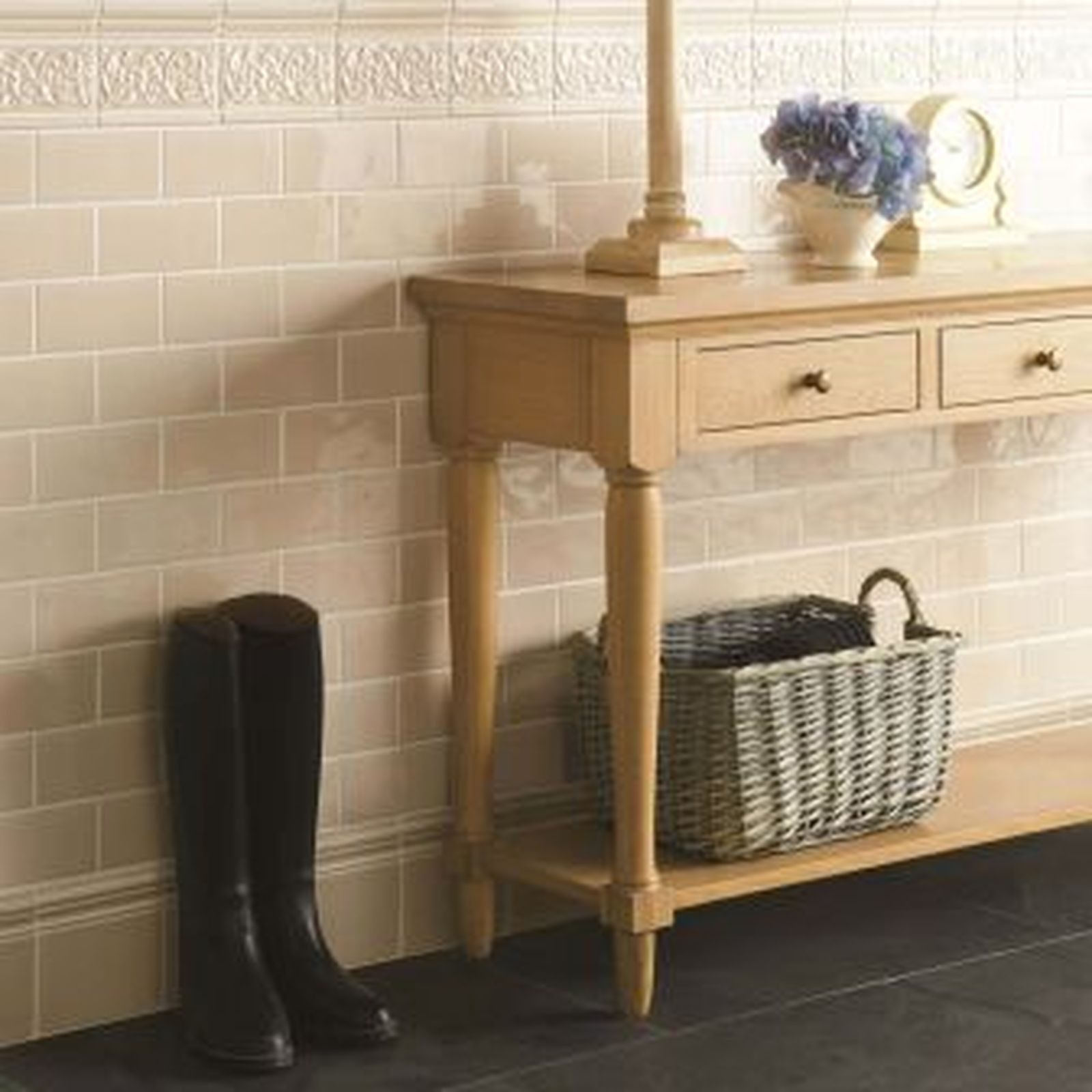 Beige tiled wall with a wooden table, basket, boots, lamp, and flowers.