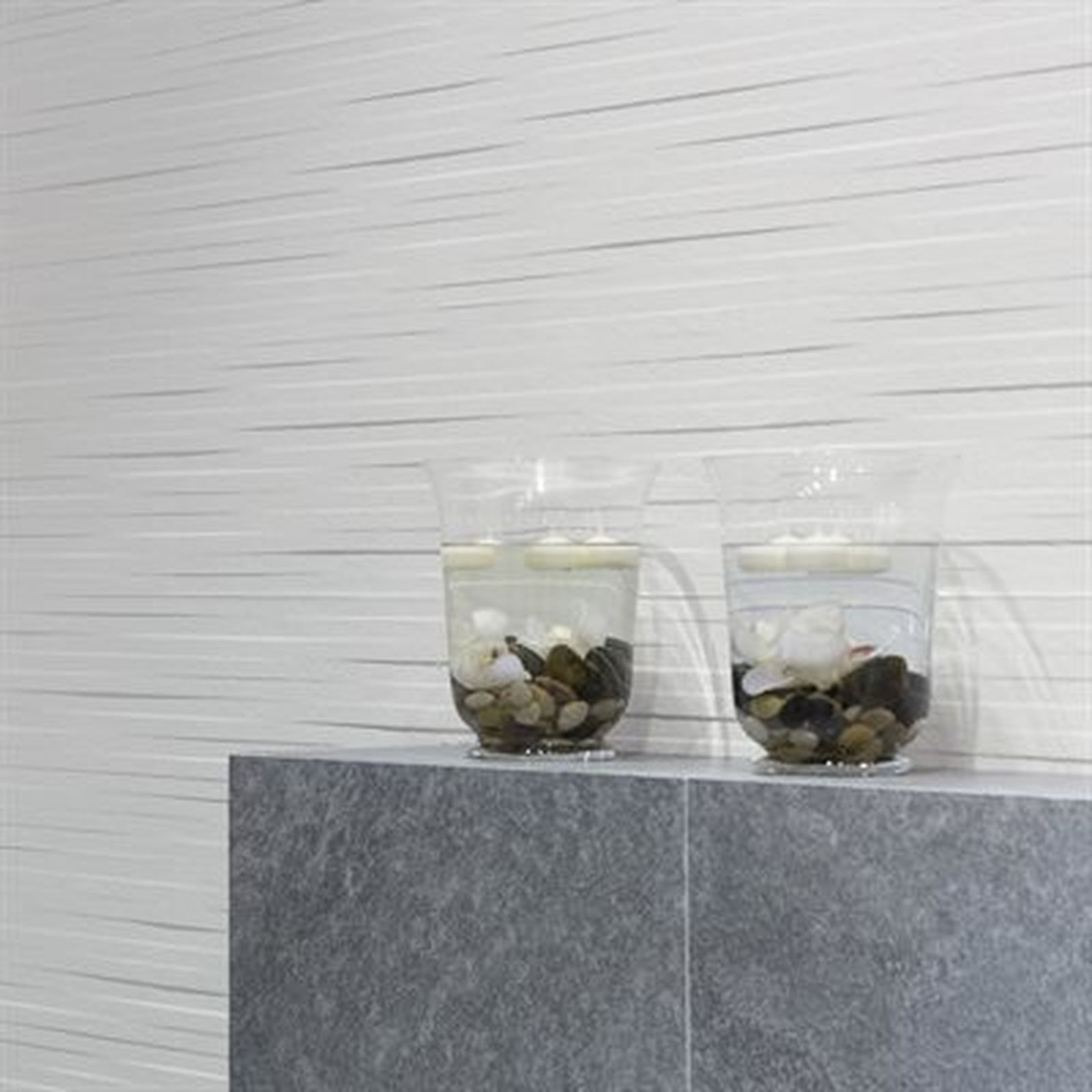 Two glasses with rocks floating in water, on gray ledge, against white ridged wall.