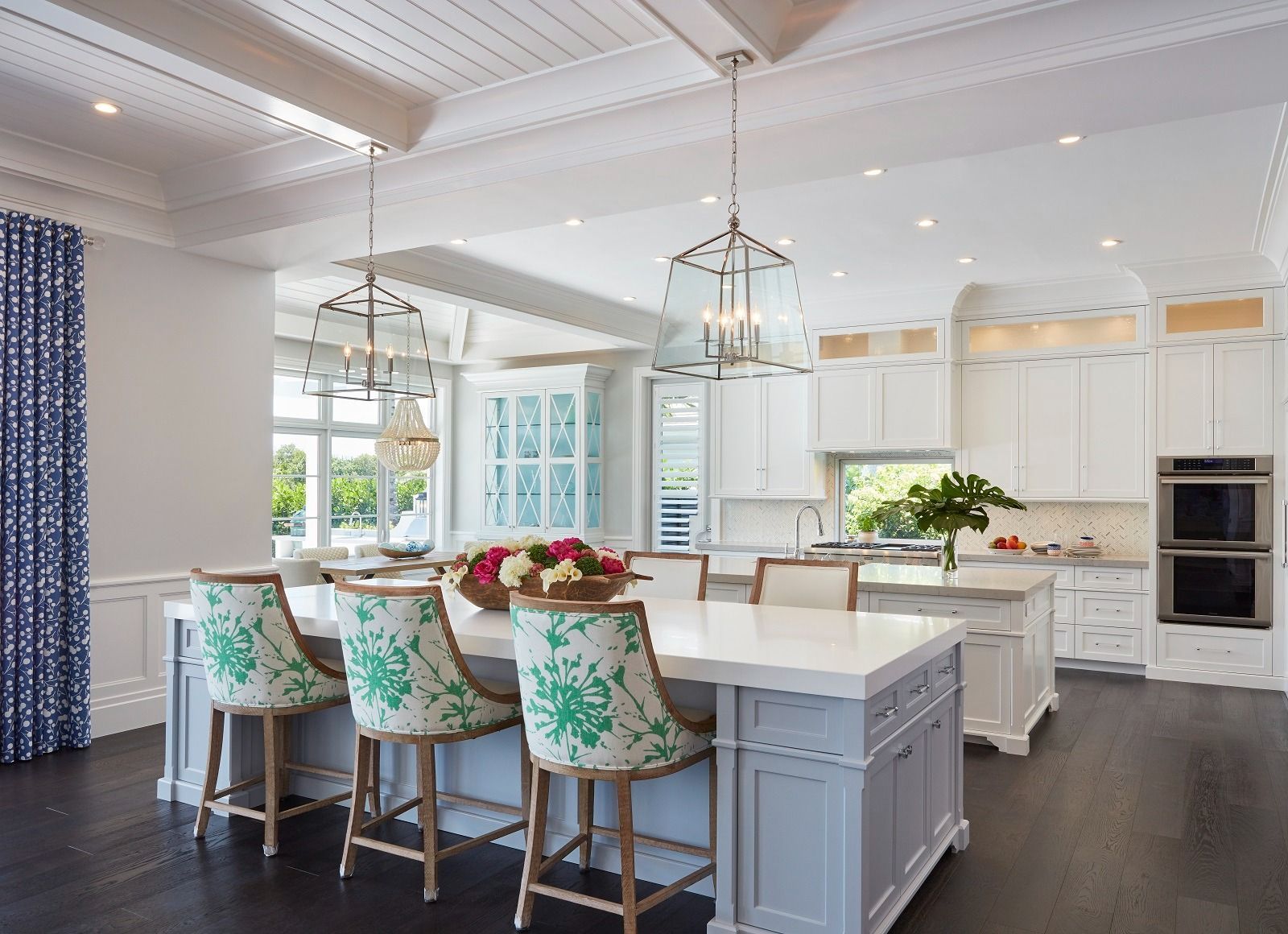 Spacious white kitchen with island, seating, and floral barstools. Dark wood floors, blue patterned drapes.