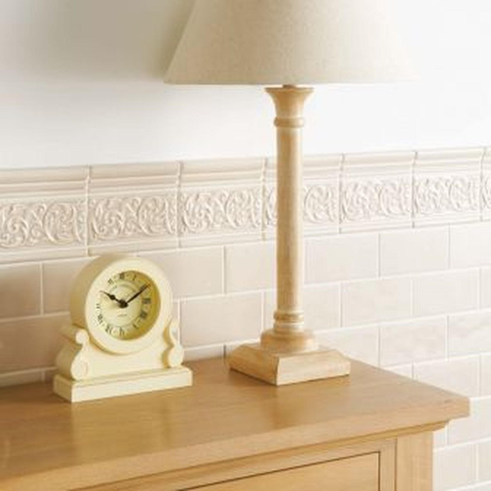 Cream-colored lamp and clock on a light wood dresser against a tile wall.
