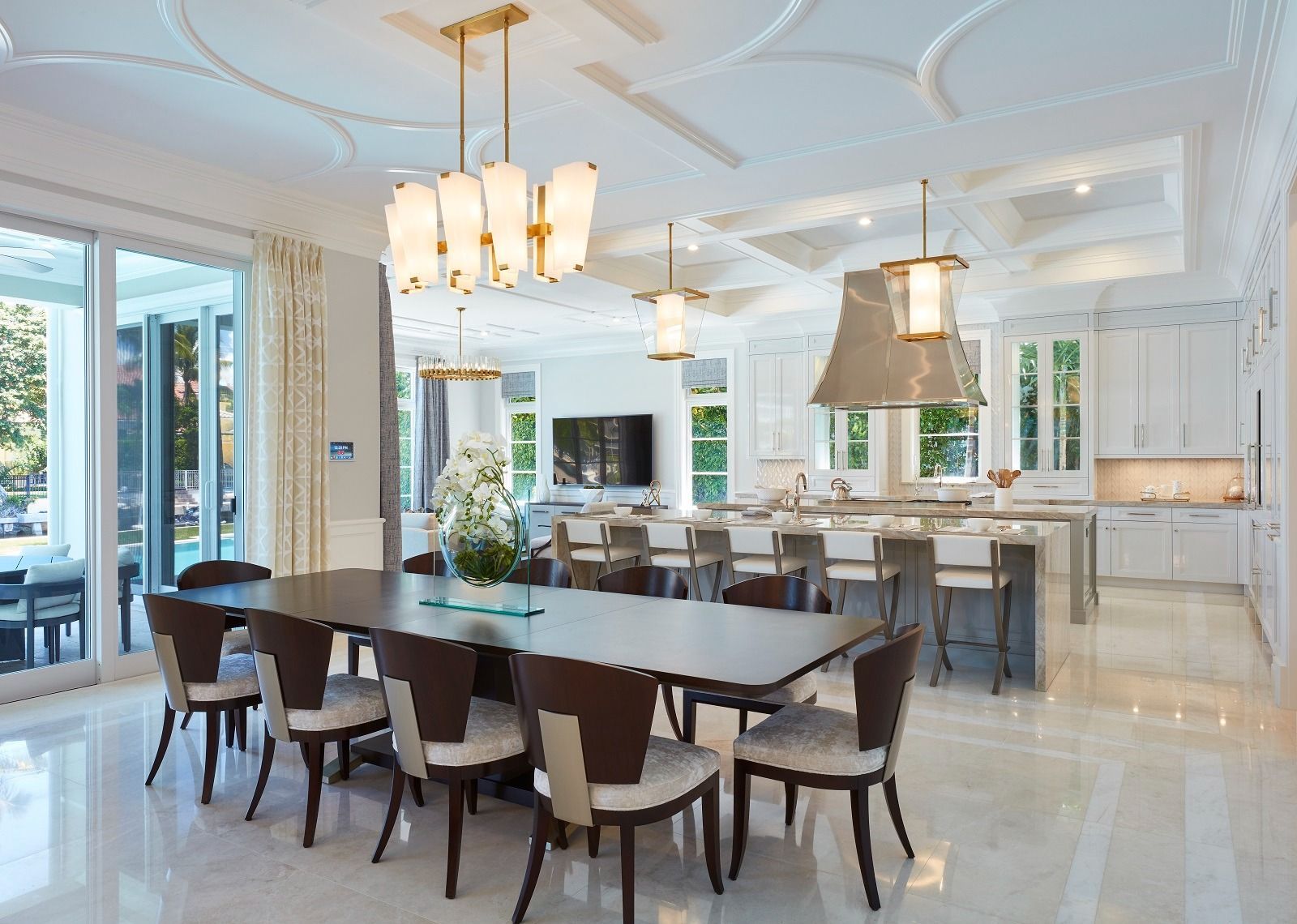 Elegant dining room with long dark table, chairs, and chandelier. Open to kitchen and outdoor patio.