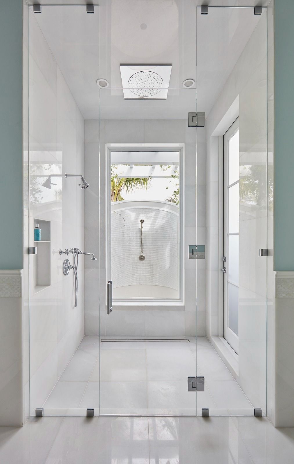 Modern glass shower enclosure in white tiled bathroom with a window.
