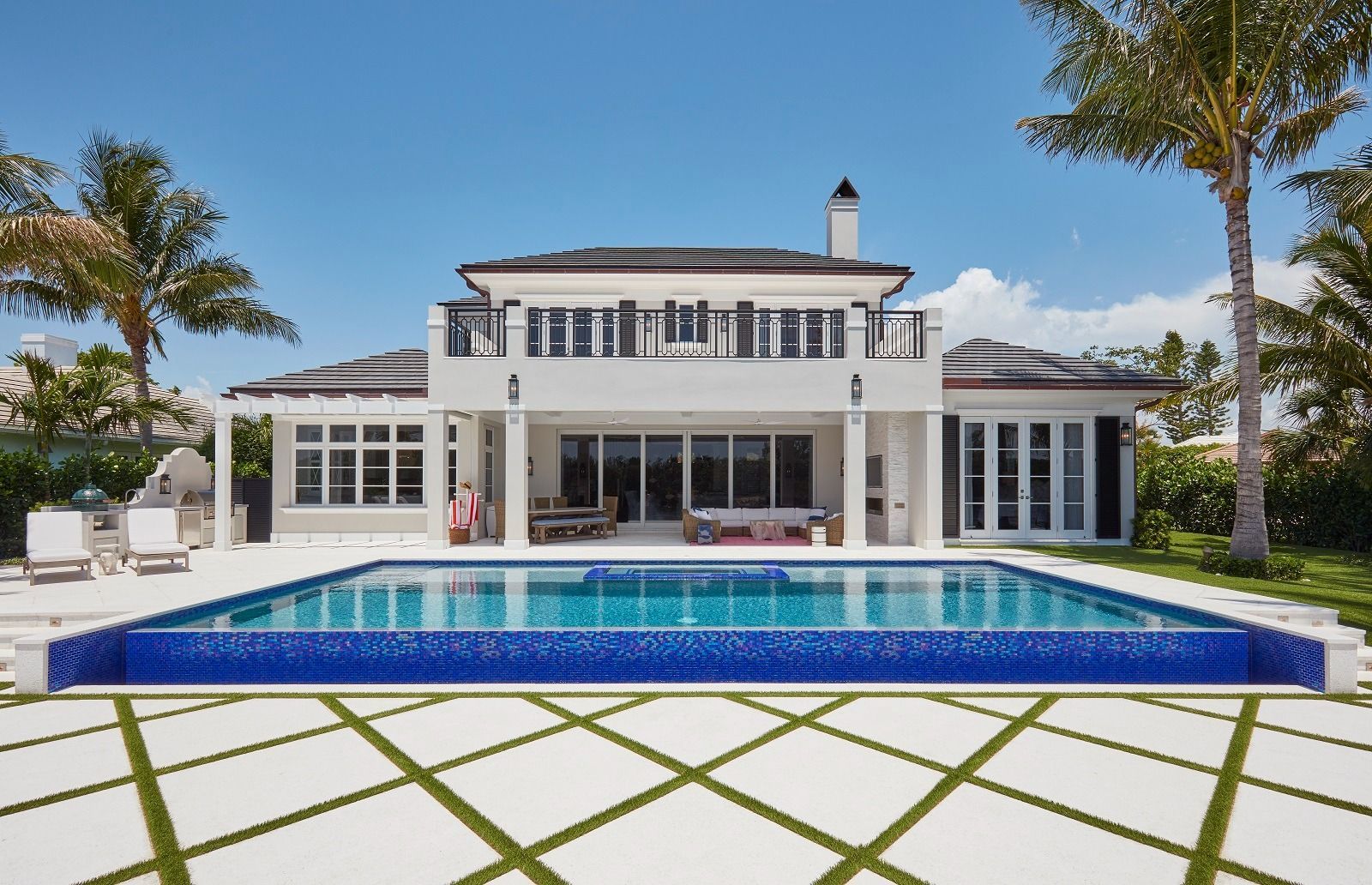 Luxury home with a blue-tiled pool, white exterior, black shutters, and palm trees under a bright blue sky.
