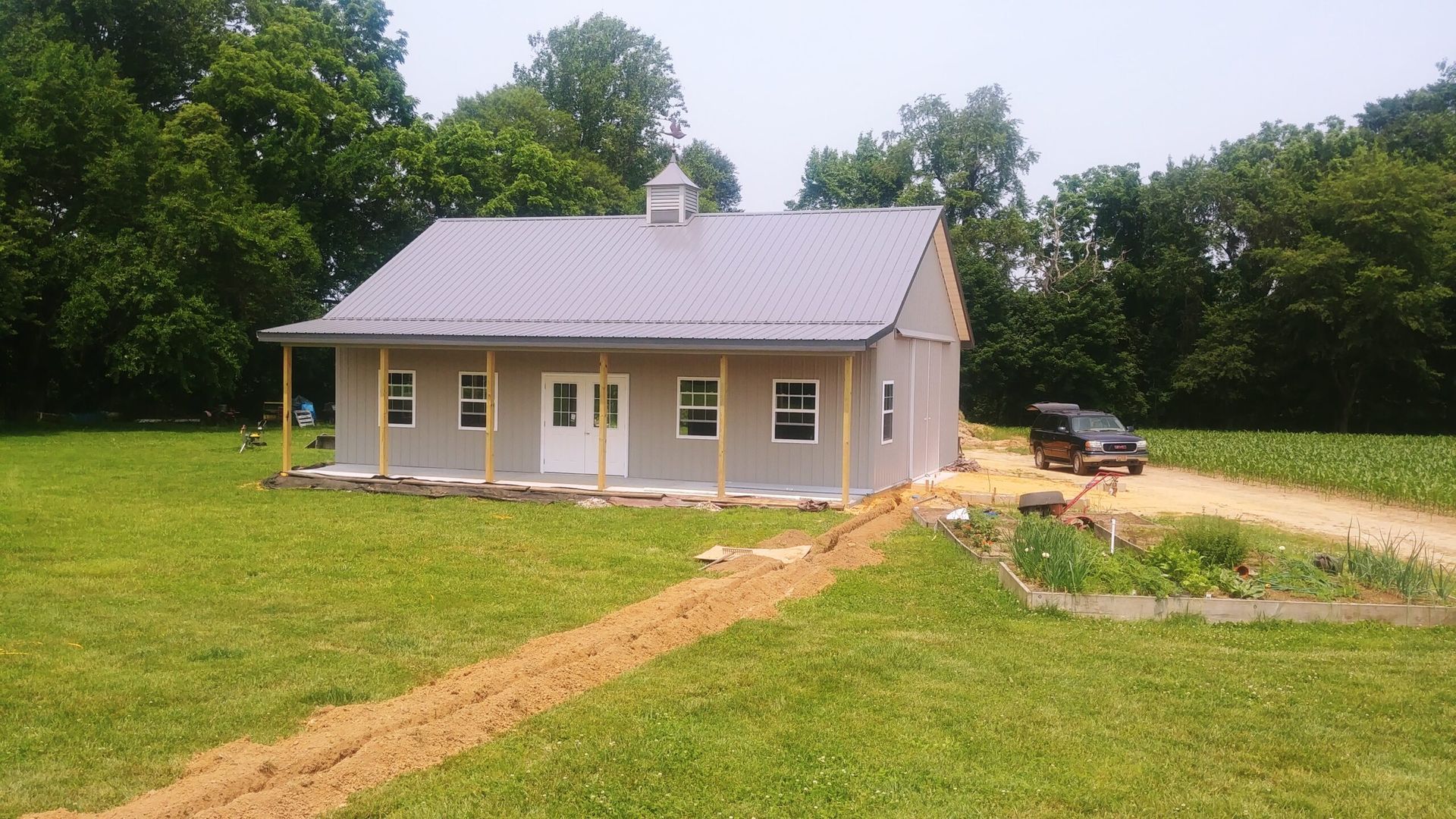 Small beige house with a metal roof on a grassy lot, surrounded by trees and a dirt driveway.
