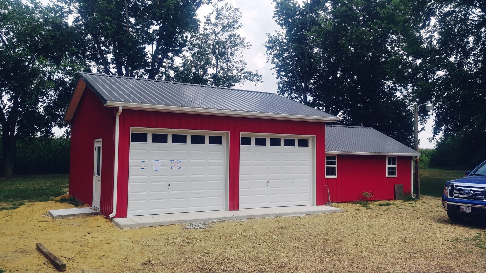 Red garage with two white doors and gray roof beside a small red shed, on a gravel driveway.